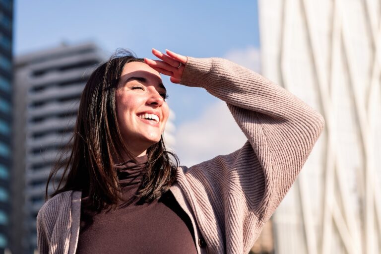 a smiling woman outside holding his hand up to cover her eyes from the sun